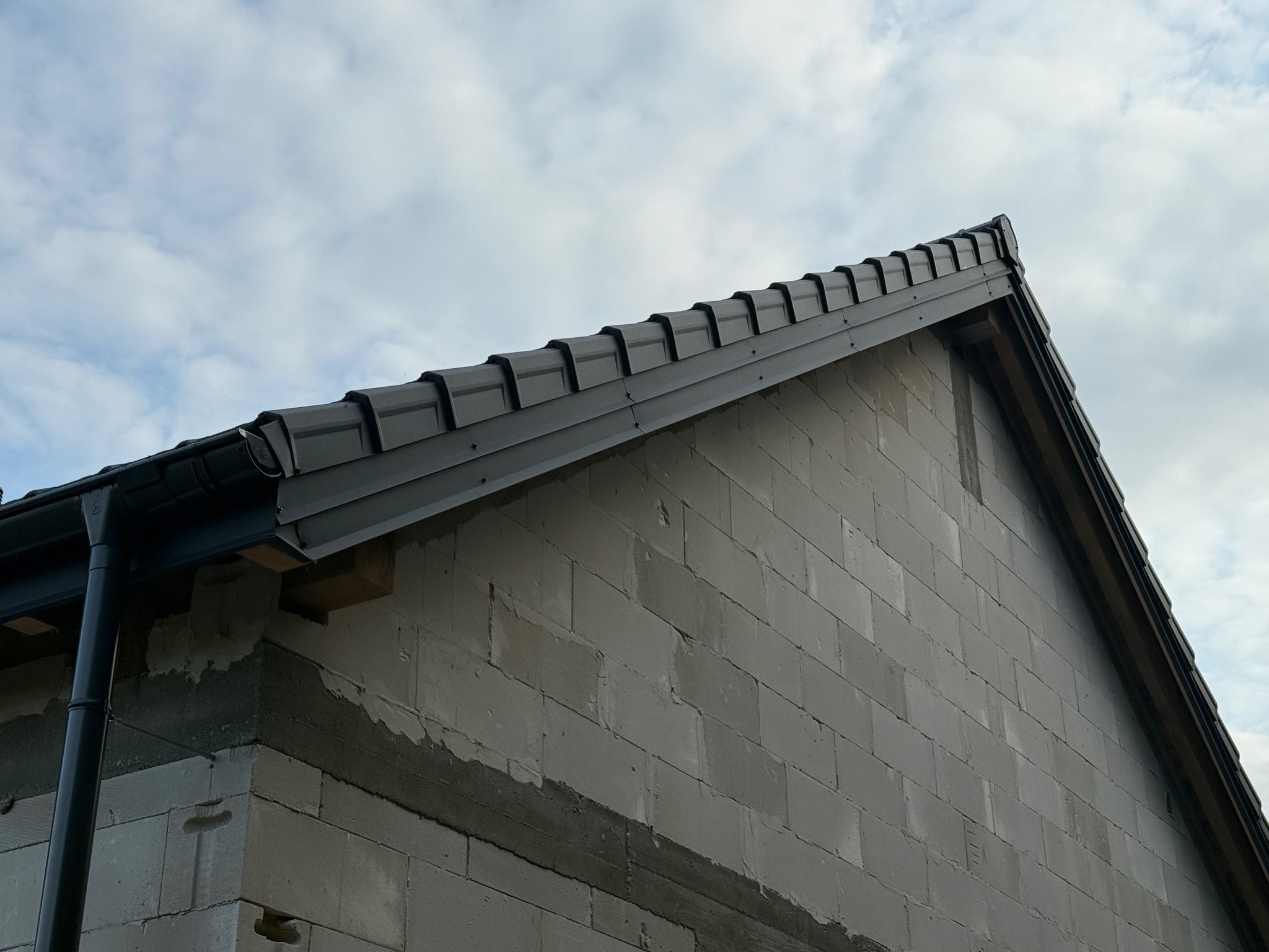 Szary dach z dachówką modułową i rynnami na surowej ścianie z betonu komórkowego, na tle pochmurnego nieba. Widok z dołu, ukośny kadr.