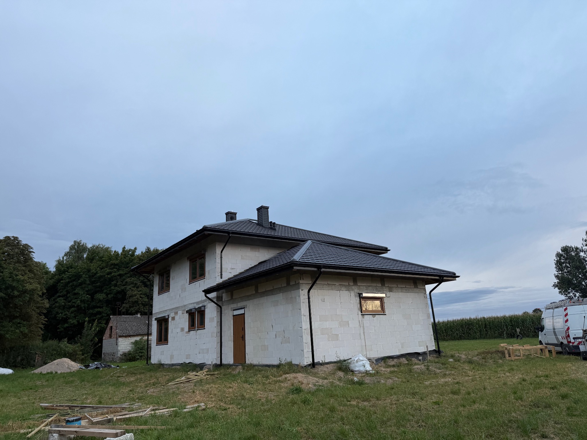 Dwukondygnacyjny dom w trakcie budowy z ciemną dachówką i rynnami. Widoczne okna z brązowymi ramami i częściowo otynkowana elewacja. W tle drzewa i niebo.