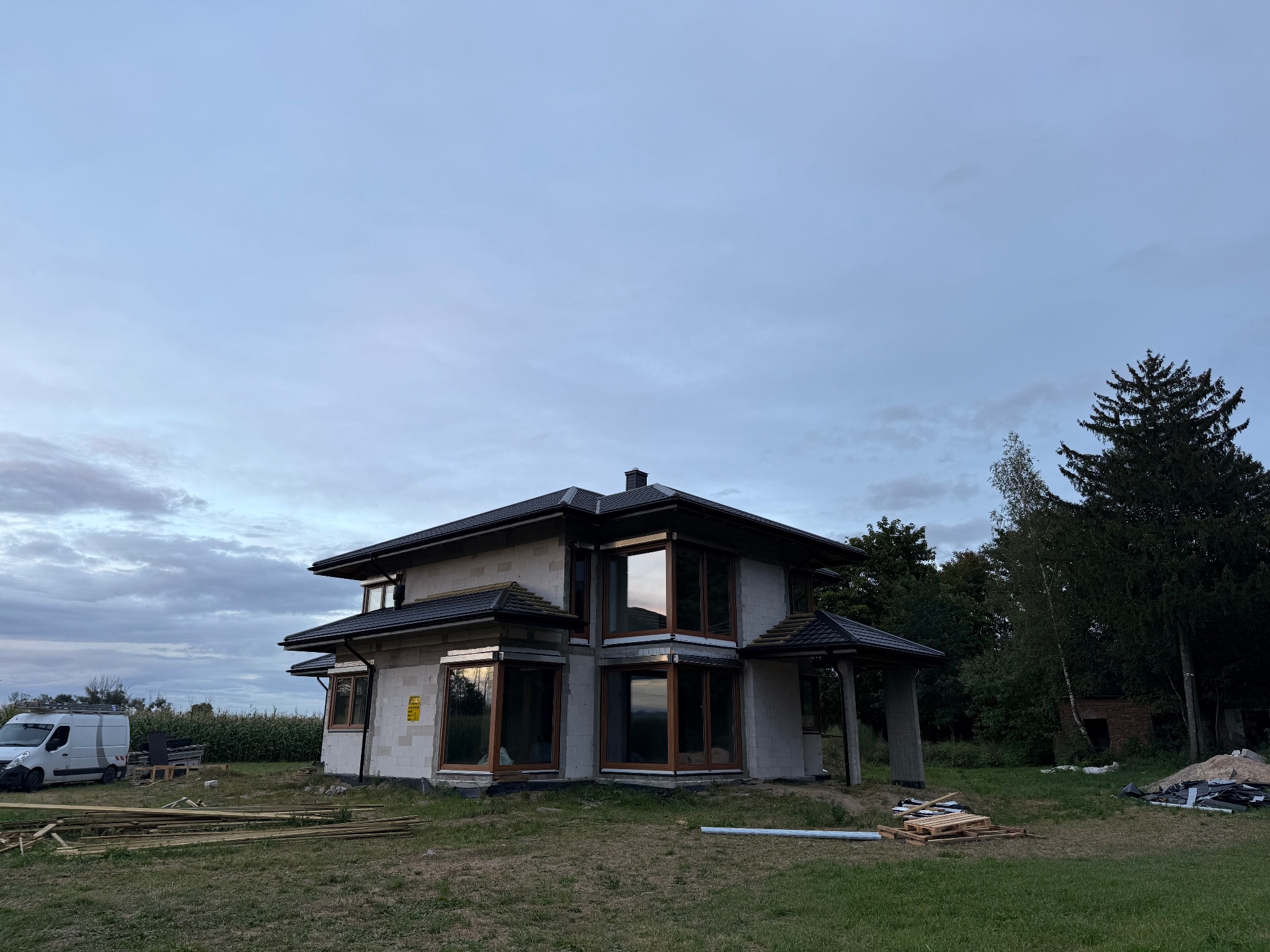 Dwukondygnacyjny dom w stanie surowym zamkniętym z ciemną dachówką, widoczny z zewnątrz na tle zachmurzonego nieba i zieleni. Częściowo widoczny samochód dostawczy.