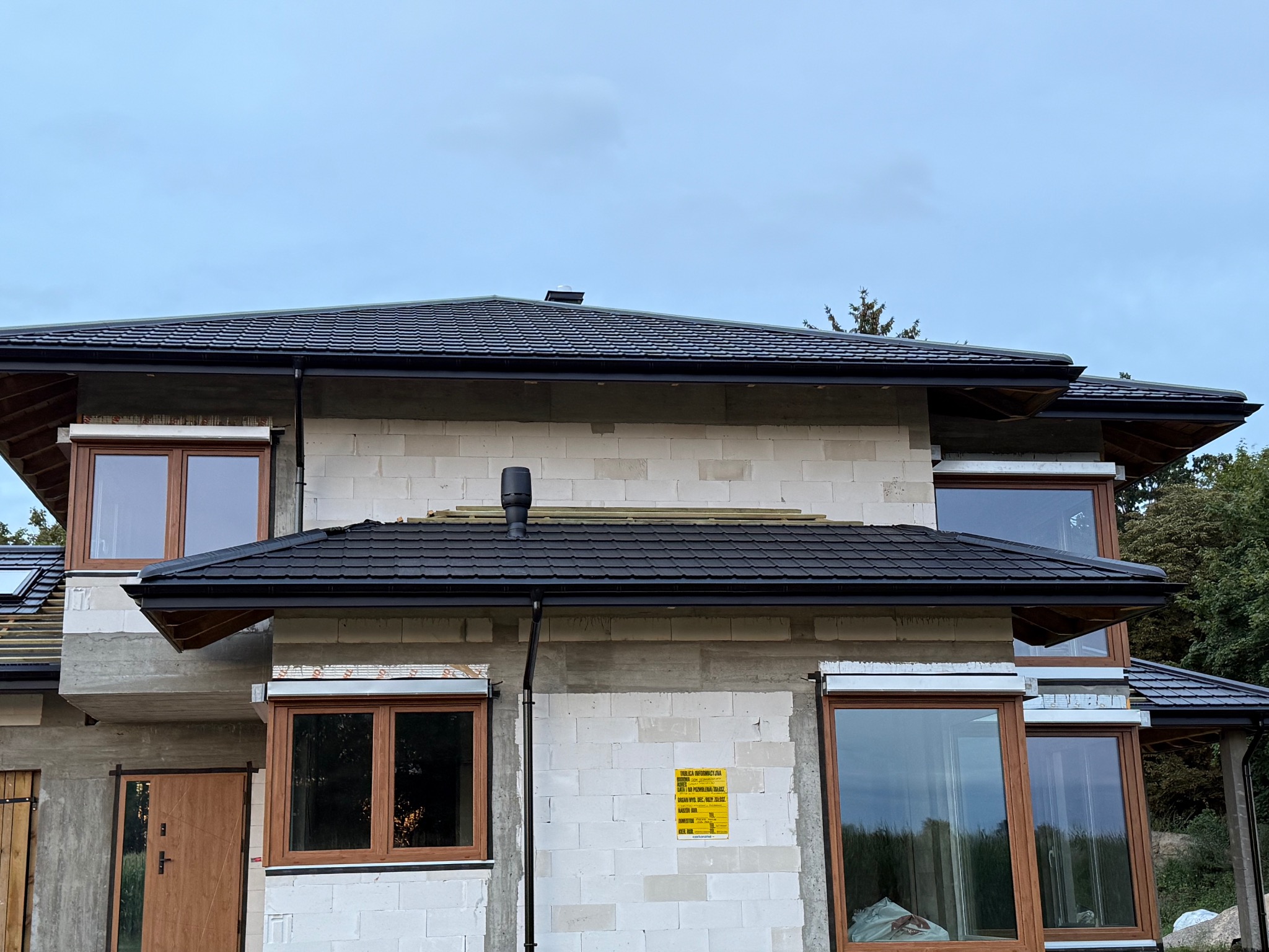 Dom w trakcie budowy z czarnym dachem z dachówki ceramicznej, widoczne okna z drewnianą stolarką i fragment niezakończonego krycia dachu po lewej stronie.