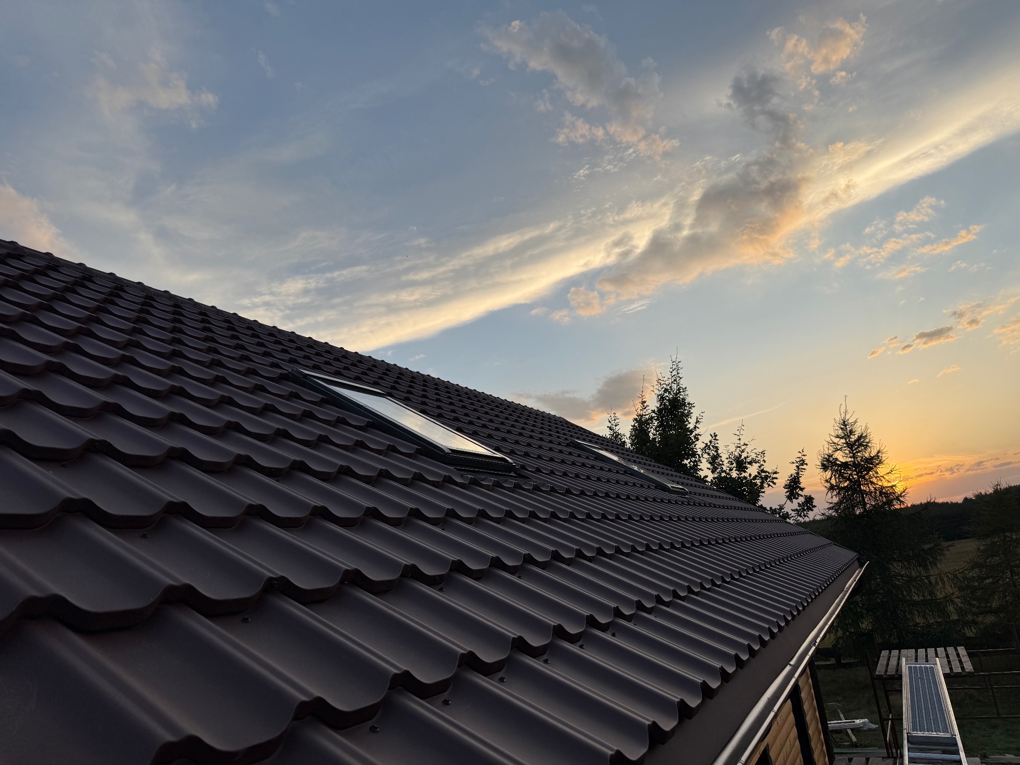 Ciemnobrązowy dach z dachówki modułowej, dwa okna dachowe, widok na zachodzące słońce i drzewa w tle. Ujęcie z dołu, lekko z prawej strony.