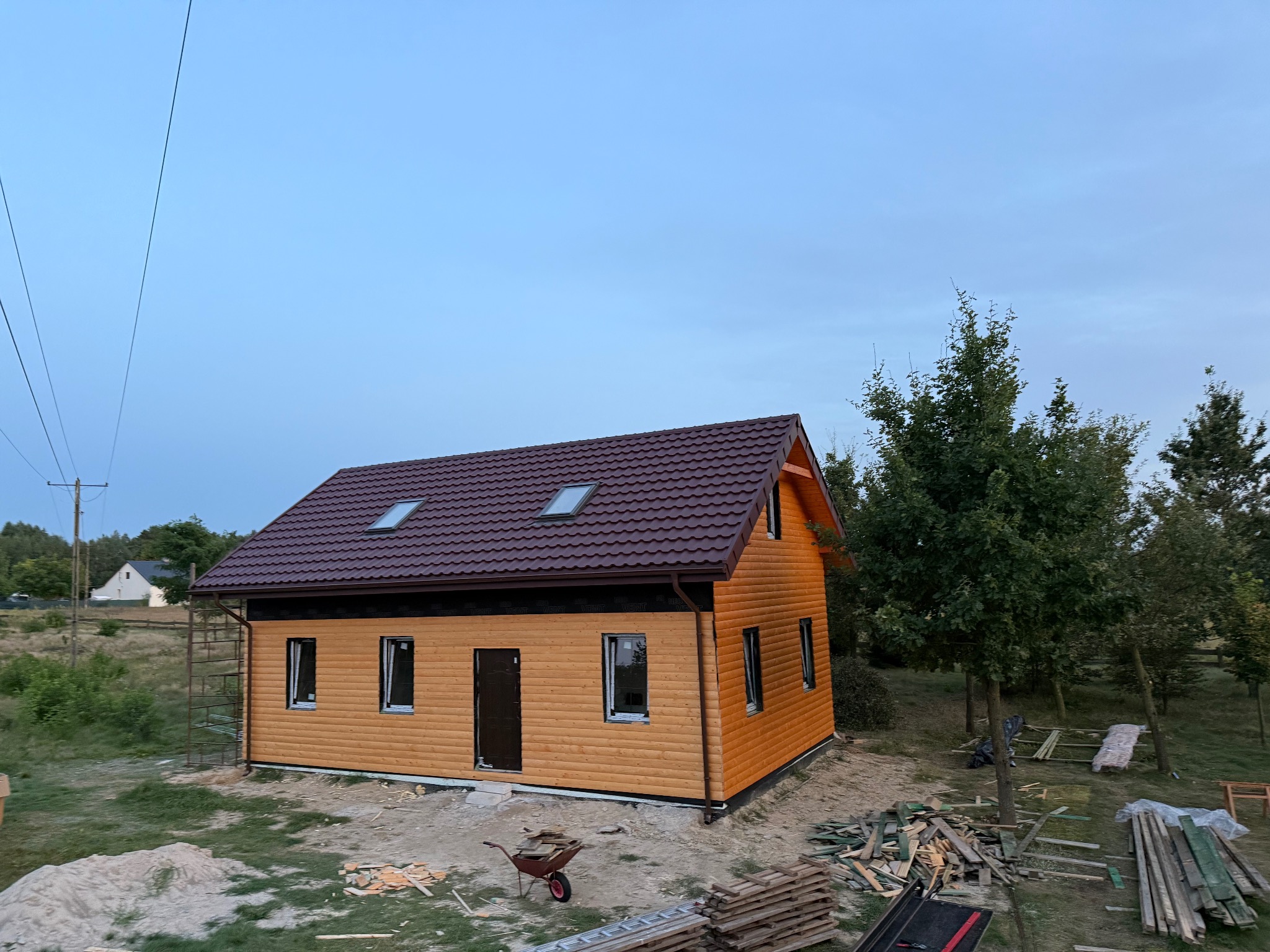 Nowy dom z brązowym dachem z dachówki i drewnianą elewacją w ciepłym odcieniu, widoczny wczesnym rankiem, z elementami rusztowania i materiałów budowlanych.