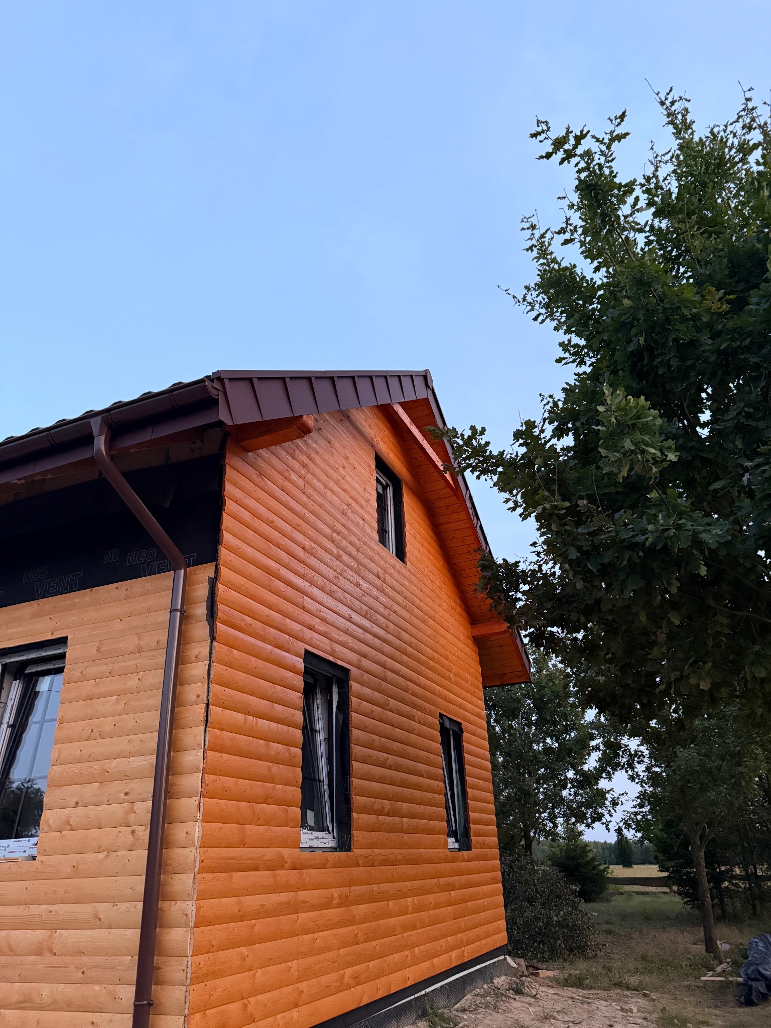 Dom z drewnianą elewacją w trakcie budowy, widoczne rynny i membrana dachowa. Ujęcie z dołu, zieleń drzew w tle.