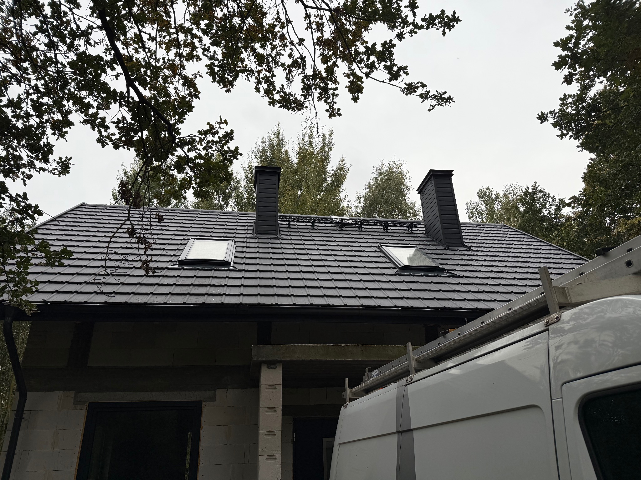Nowy, grafitowy dach z dwoma oknami dachowymi i kominami na surowym budynku. Na dachu widoczna drabina oparta o samochód dostawczy. Ujęcie z dołu, częściowo zasłonięte przez drzewa.