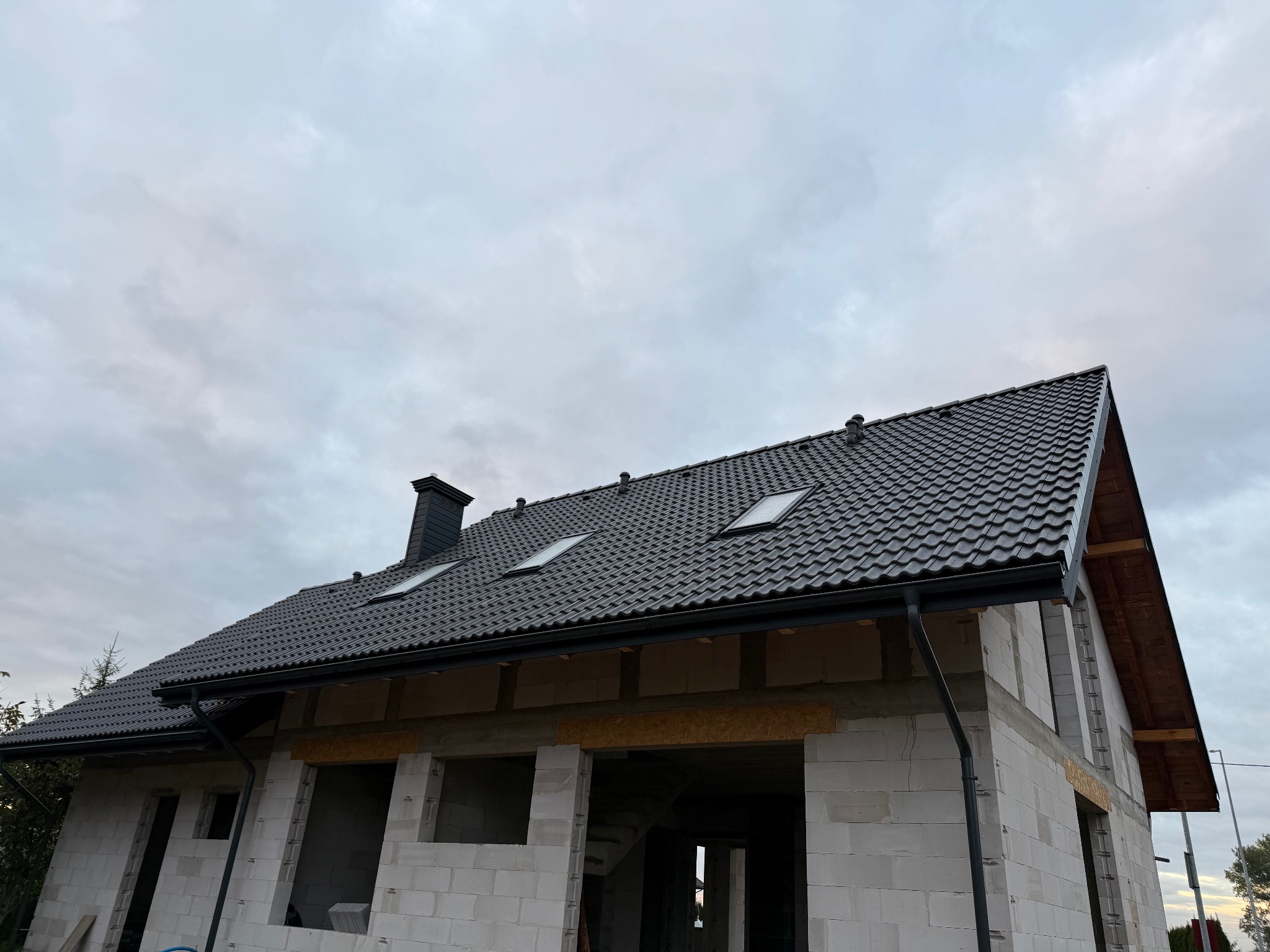 Nowy, grafitowy dach z dachówką ceramiczną i oknami połaciowymi na surowym budynku z białego betonu komórkowego, widok z dołu na tle pochmurnego nieba.