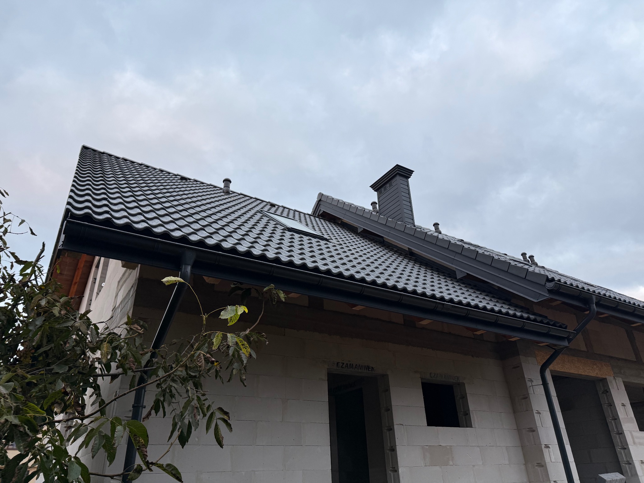 Grafitowy dach z dachówki ceramicznej na surowym budynku, widok z dołu, z rynnami i kominem. Częściowo widoczne drzewo z liśćmi. Pochmurne niebo w tle.
