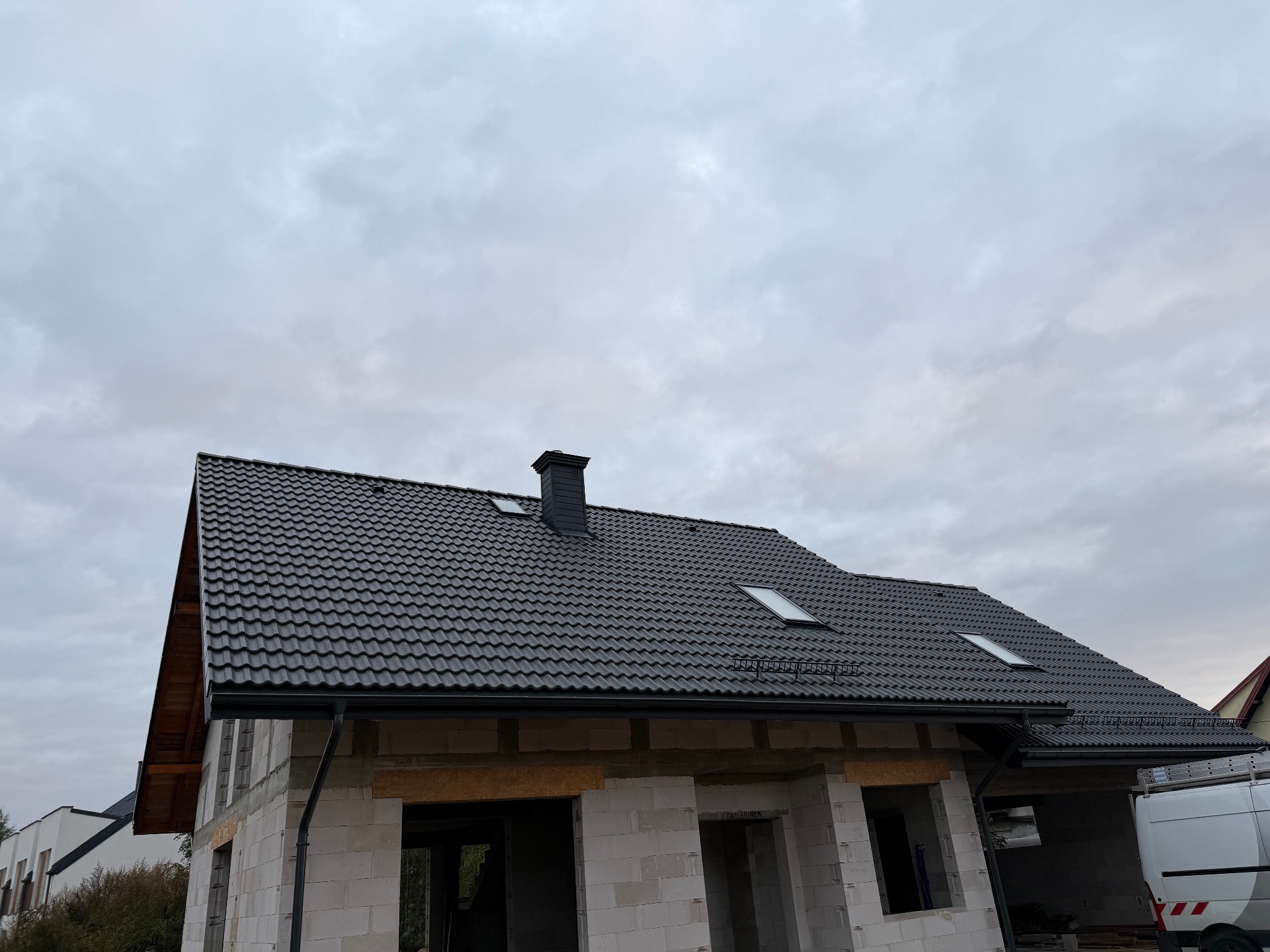 Dom w trakcie budowy z ciemną dachówką, widoczne okna dachowe i komin. Na pierwszym planie białe bloczki konstrukcyjne, w tle pochmurne niebo.