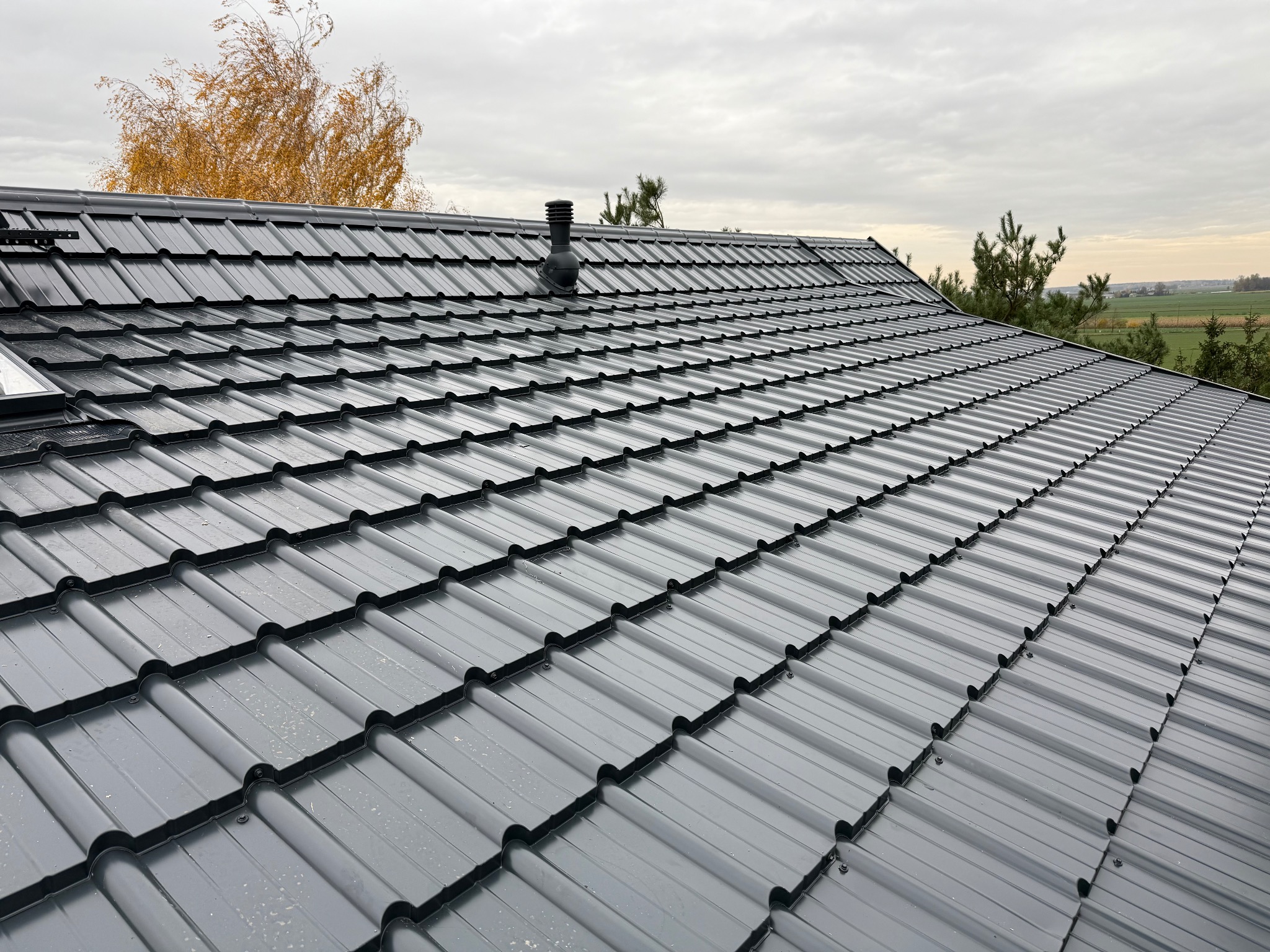 Nowoczesny, grafitowy dach z dachówki modułowej, z kominkiem wentylacyjnym i oknem dachowym, na tle jesiennego krajobrazu i pochmurnego nieba.