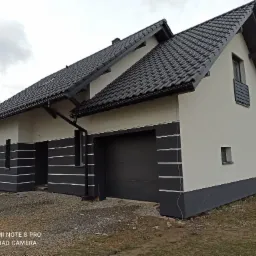 Nowoczesny dom jednorodzinny z białą elewacją i ciemnoszarymi detalami, widoczne wykończenie z poziomymi paskami, grafitowa dachówka i brama garażowa, teren wokół domu w trakcie zagospodarowania.