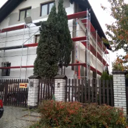 Dom jednorodzinny w trakcie ocieplania, widoczne rusztowania, styropianowe płyty izolacyjne na ścianach, częściowo otynkowana elewacja, drewniany płot z ceglanymi słupkami i roślinnością przed...