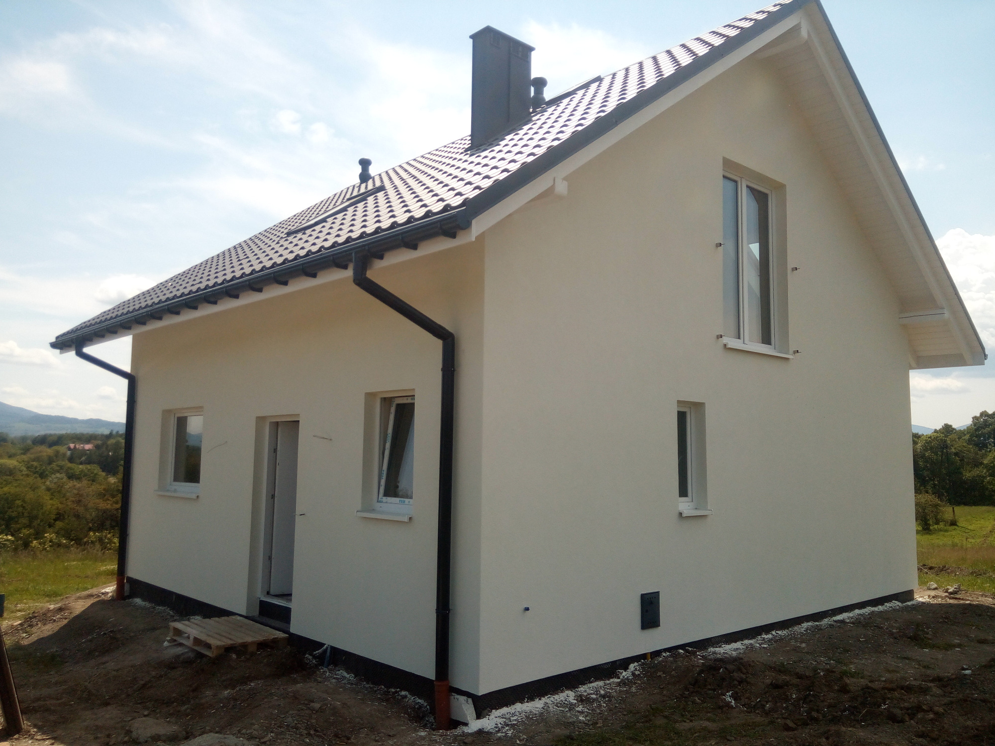 Nowoczesny dom jednorodzinny z jasną elewacją, ciemną dachówką i czarnymi rynnami, widok z zewnątrz na tle zieleni i gór.