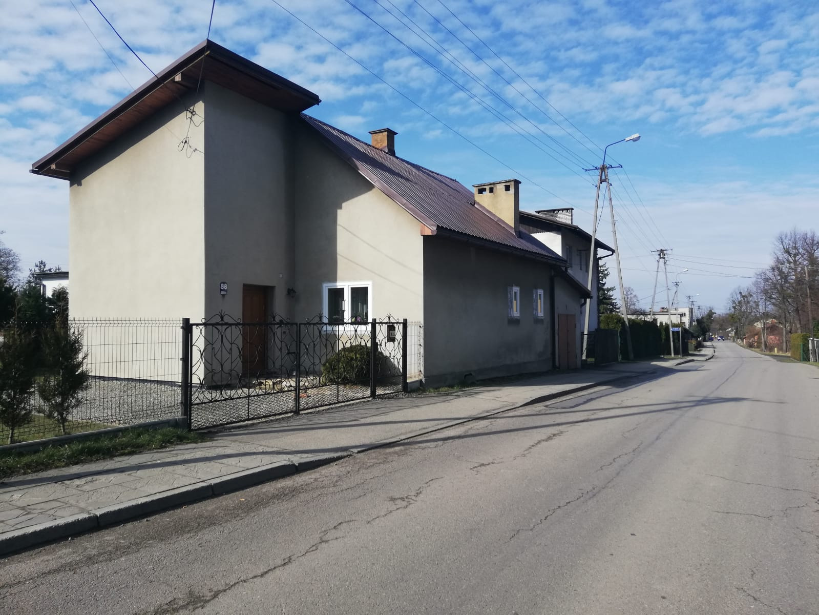 Dwupiętrowy dom z szarą elewacją i brązowym dachem, widziany z ulicy w słoneczny dzień. Ozdobne metalowe ogrodzenie z bramą przed domem. Widoczne słupy energetyczne i błękitne niebo z chmurami.