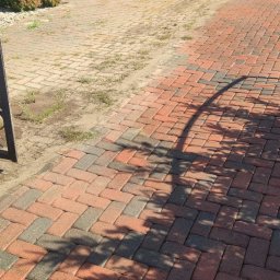 ZACZYSZCZONE PIOTR STĘPNIAK - Ułożona kostka brukowa w odcieniach czerwieni i szarości, częściowo porośnięta trawą, z cieniem drzewa i fragmentem metalowego ogrodzenia po lewej stronie.