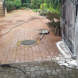 ZACZYSZCZONE PIOTR STĘPNIAK - Czerwona kostka brukowa i fragment muru z bramą w trakcie czyszczenia pianą aktywną, widoczny sprzęt do mycia ciśnieniowego i kabel zasilający.
