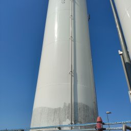 ZACZYSZCZONE PIOTR STĘPNIAK - Biały, przemysłowy zbiornik z widocznymi śladami zabrudzeń i zacieków na dolnej części, sfotografowany pod kątem od dołu, na tle błękitnego nieba.