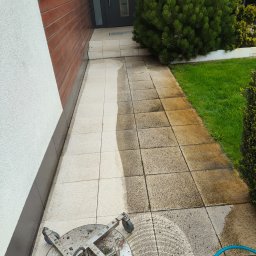 ZACZYSZCZONE PIOTR STĘPNIAK - Częściowo wyczyszczona kostka brukowa przed domem, widoczna różnica między czystą a brudną powierzchnią, obok leży urządzenie do czyszczenia ciśnieniowego z niebieskim wężem.