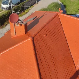 Widok z góry na dach pokryty czerwoną dachówką bitumiczną w kształcie rybiej łuski, z ceglanym kominem, anteną satelitarną i ozdobnym kogutem na kalenicy.