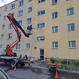 ZACZYSZCZONE PIOTR STĘPNIAK - Pracownik na podnośniku czyści elewację bloku, widoczny efekt różnicy czystości. Na dole agregat ciśnieniowy, rower i zaparkowany samochód. Ujęcie z dołu.