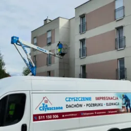 Pracownik na podnośniku koszowym czyści elewację budynku. Na pierwszym planie biały bus z reklamą firmy oferującej usługi czyszczenia.