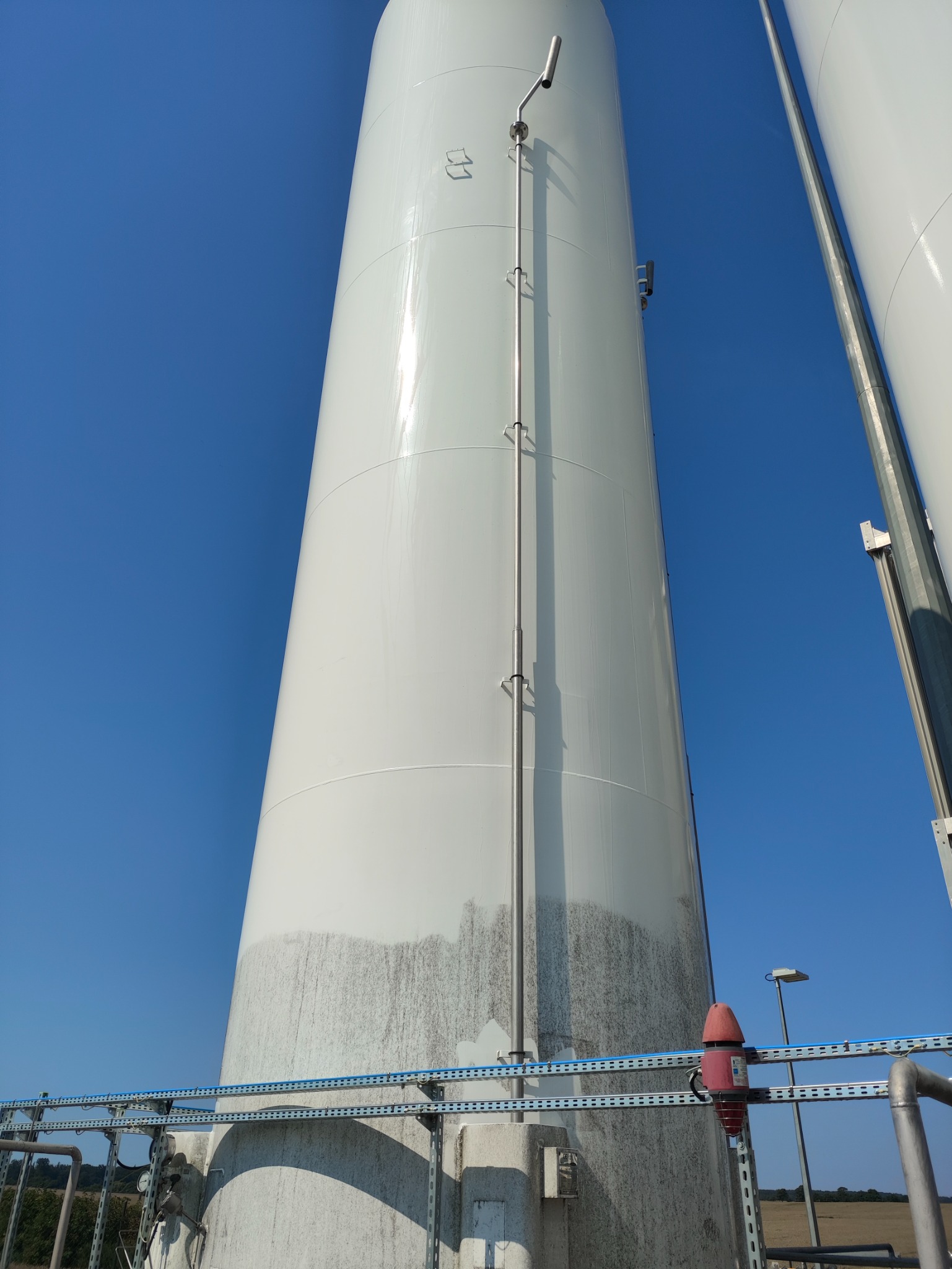 Biały, przemysłowy zbiornik z widocznymi śladami zabrudzeń i zacieków na dolnej części, sfotografowany pod kątem od dołu, na tle błękitnego nieba.