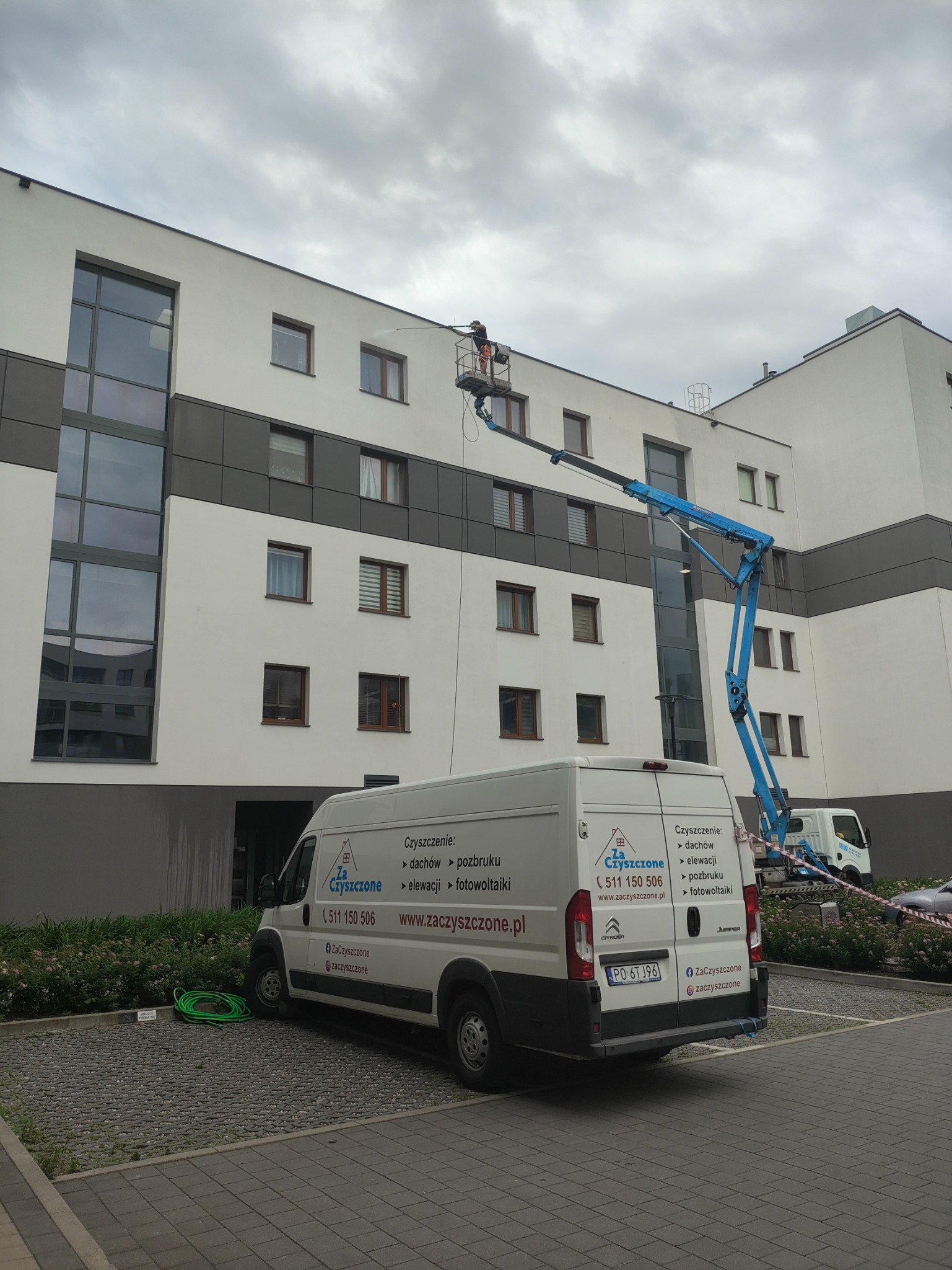 Pracownik na podnośniku koszowym czyści elewację budynku, widoczny biały bus z logo firmy świadczącej usługi czyszczenia, w tle pochmurne niebo.