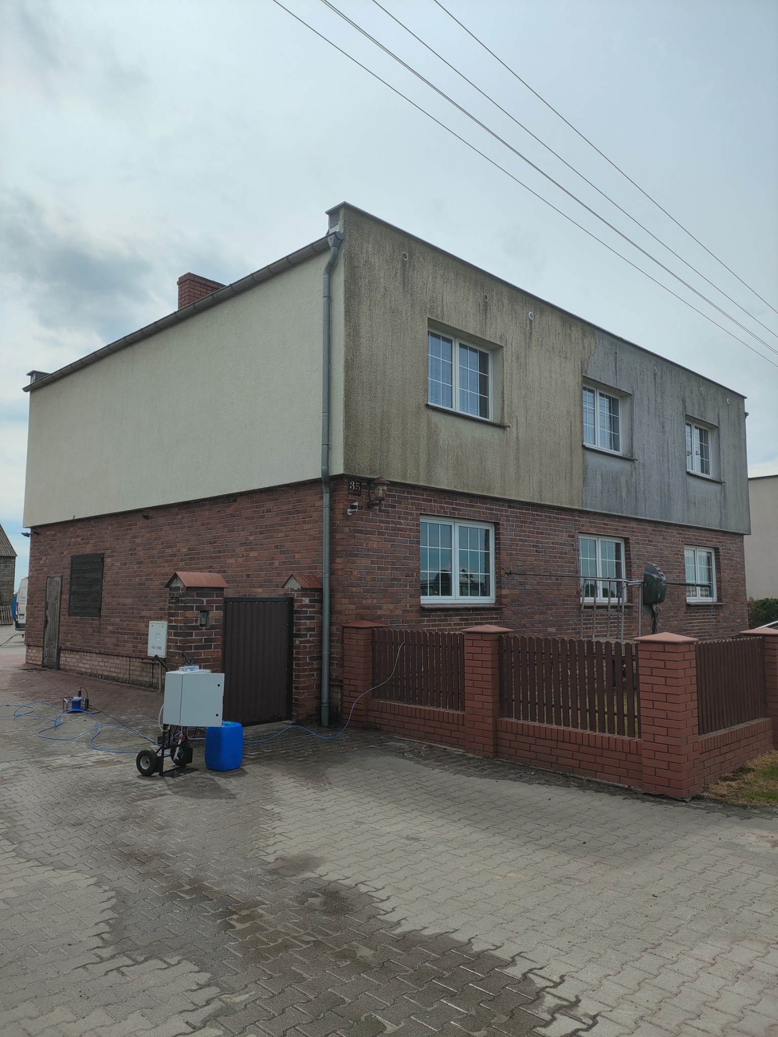 Dwukondygnacyjny dom w Tarnowie Podgórnym z ceglaną dolną częścią i jasną górną, z widocznymi śladami zabrudzeń na elewacji. Na pierwszym planie urządzenie do czyszczenia pod ciśnieniem i niebieski...