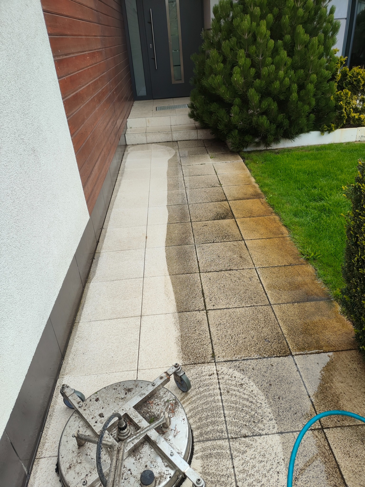 Częściowo wyczyszczona kostka brukowa przed domem, widoczna różnica między czystą a brudną powierzchnią, obok leży urządzenie do czyszczenia ciśnieniowego z niebieskim wężem.