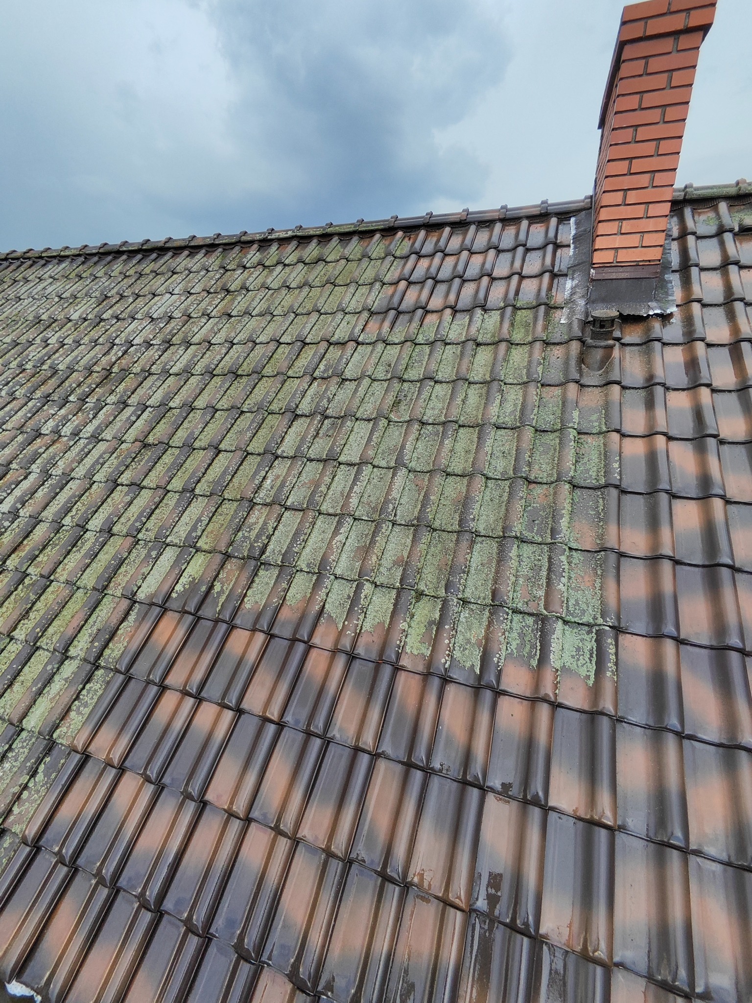Częściowo oczyszczony dach z ceglastą dachówką, widoczny komin z cegły i fragment pochmurnego nieba.
