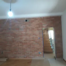 Pokój w trakcie prac wykończeniowych z czerwoną cegłą na ścianie, widoczne zabezpieczenia podłogi i przygotowane otwory na gniazdka elektryczne.