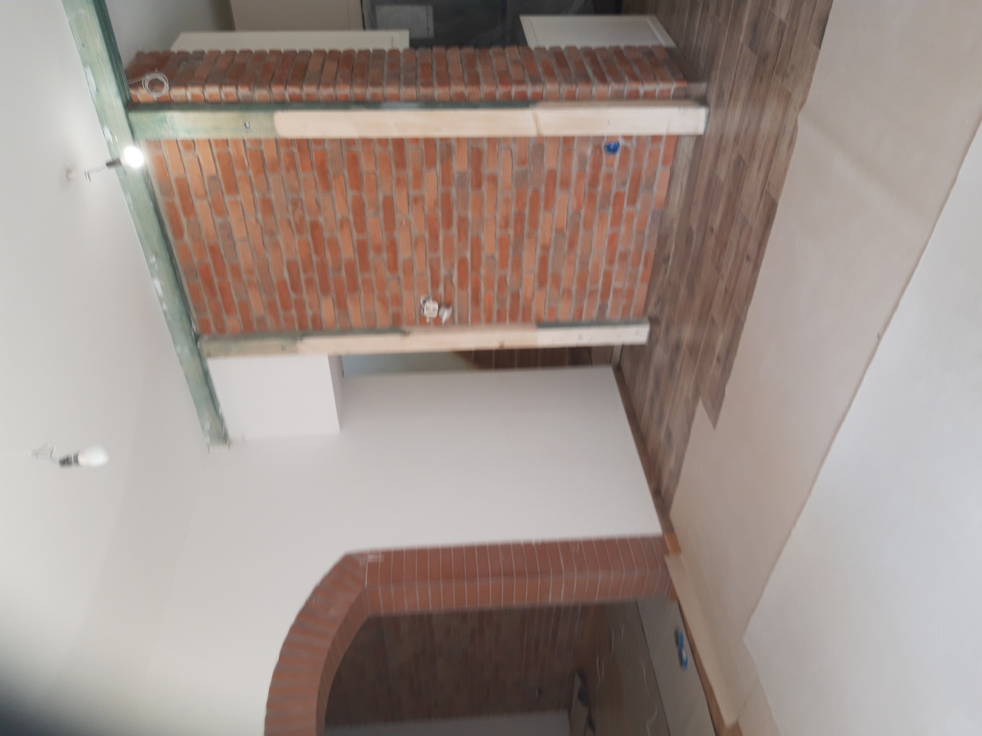 Wnętrze w trakcie prac wykończeniowych: fragment ściany z cegły klinkierowej, drewniane elementy konstrukcyjne, fragment łuku z cegły, panele podłogowe i białe ściany.