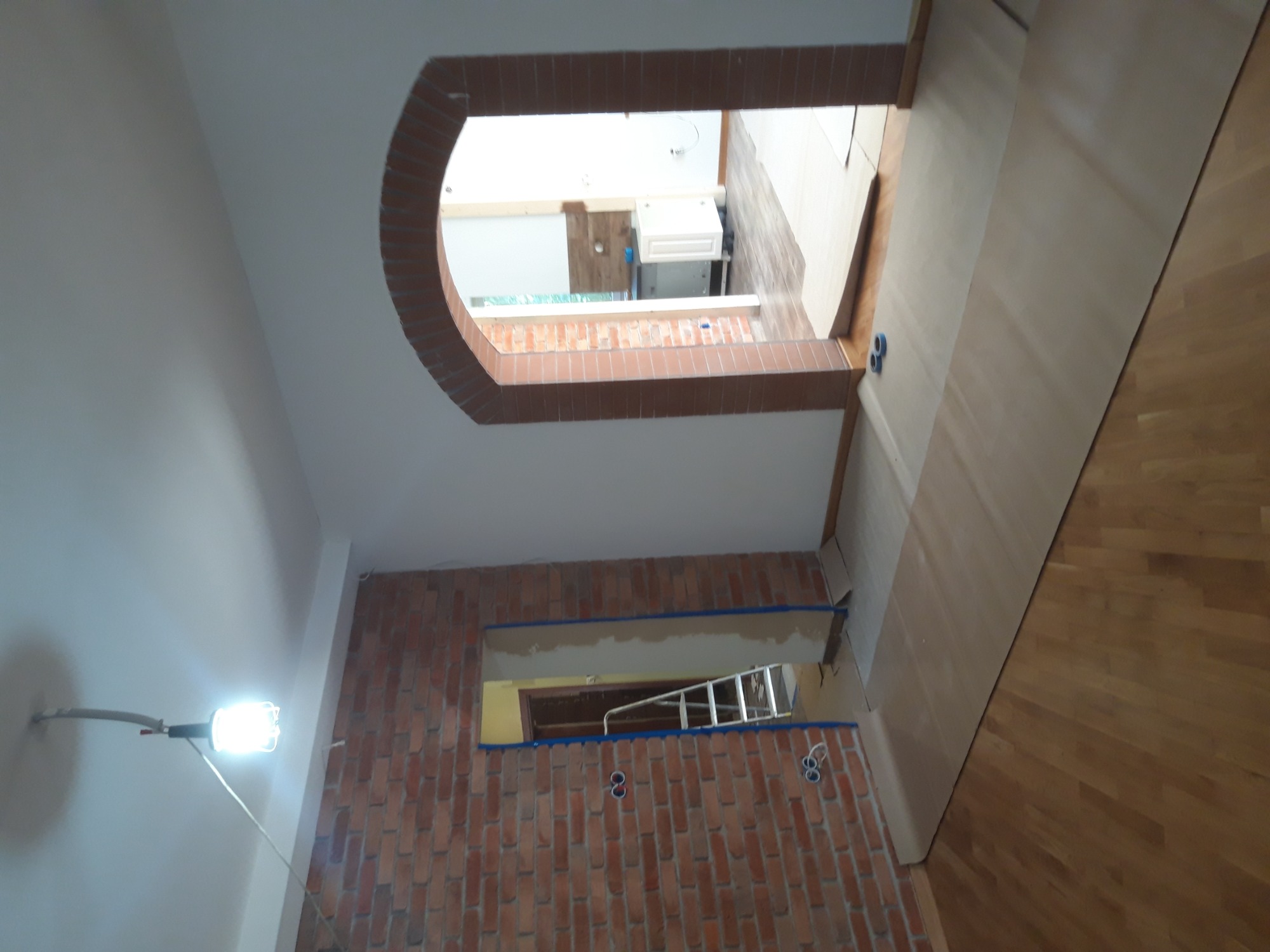 Wnętrze w trakcie prac wykończeniowych: fragment ściany z cegły klinkierowej, zabezpieczona taśmą malarską, widoczny fragment sufitu z okrągłym przejściem, podłoga zabezpieczona kartonem, drabina...