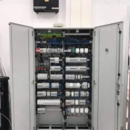 Otwarta szafa sterownicza z licznymi przekaźnikami, bezpiecznikami i okablowaniem, zamontowana w pomieszczeniu z białą ścianą, nad szafą widoczna skrzynka LKD z symbolem ostrzegawczym.