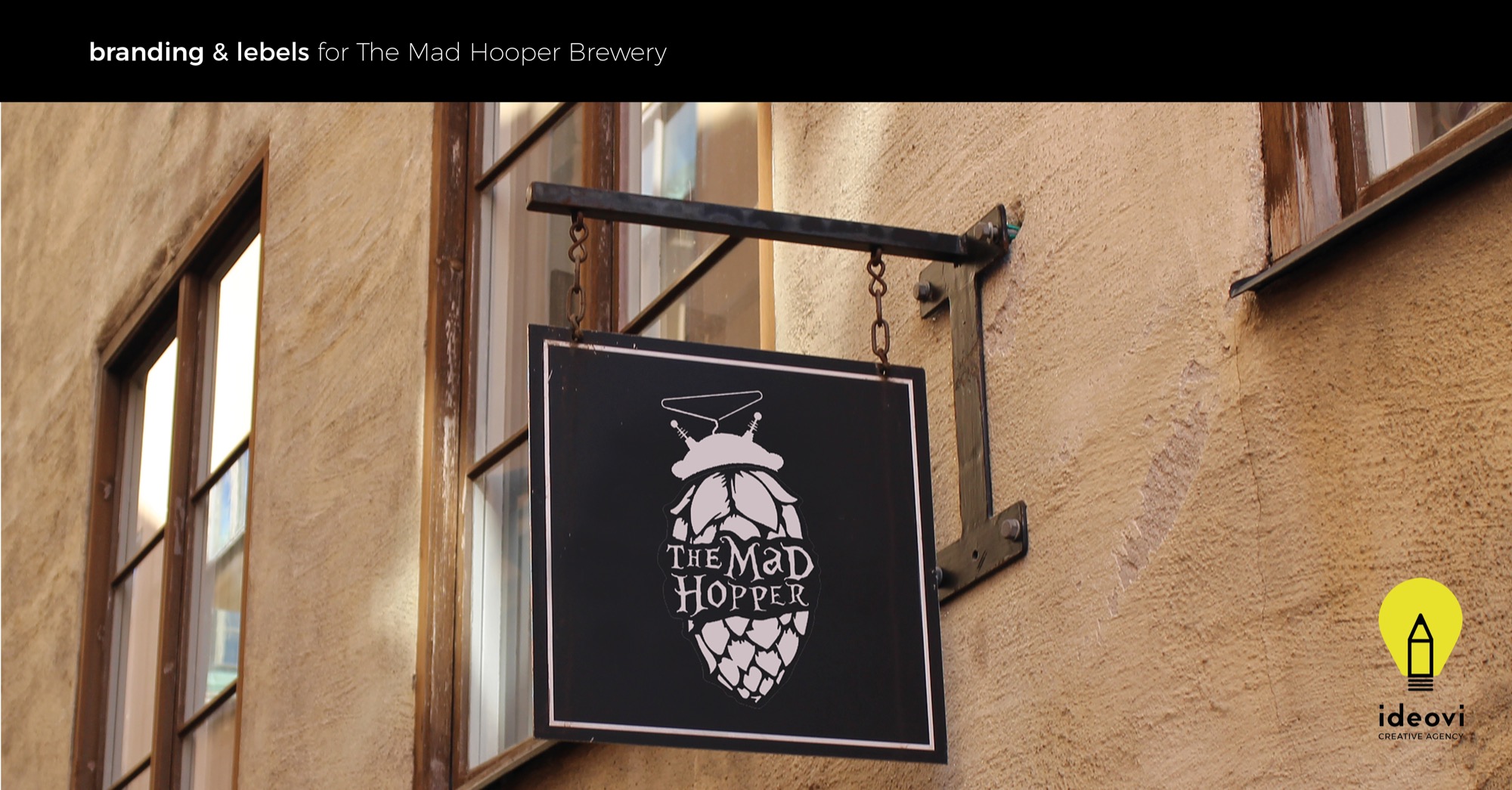 Znak firmowy browaru The Mad Hooper z motywem chmielu, zawieszony na ścianie budynku z oknami.