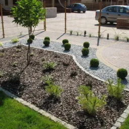 Kwadratowa rabata z korą, młodymi iglakami, topiarami z bukszpanu i białymi kamieniami, obrzeże z kostki granitowej, szara strefa z kamyczków, ścieżka z kostki brukowej w tle, w tle ogrodzenie...