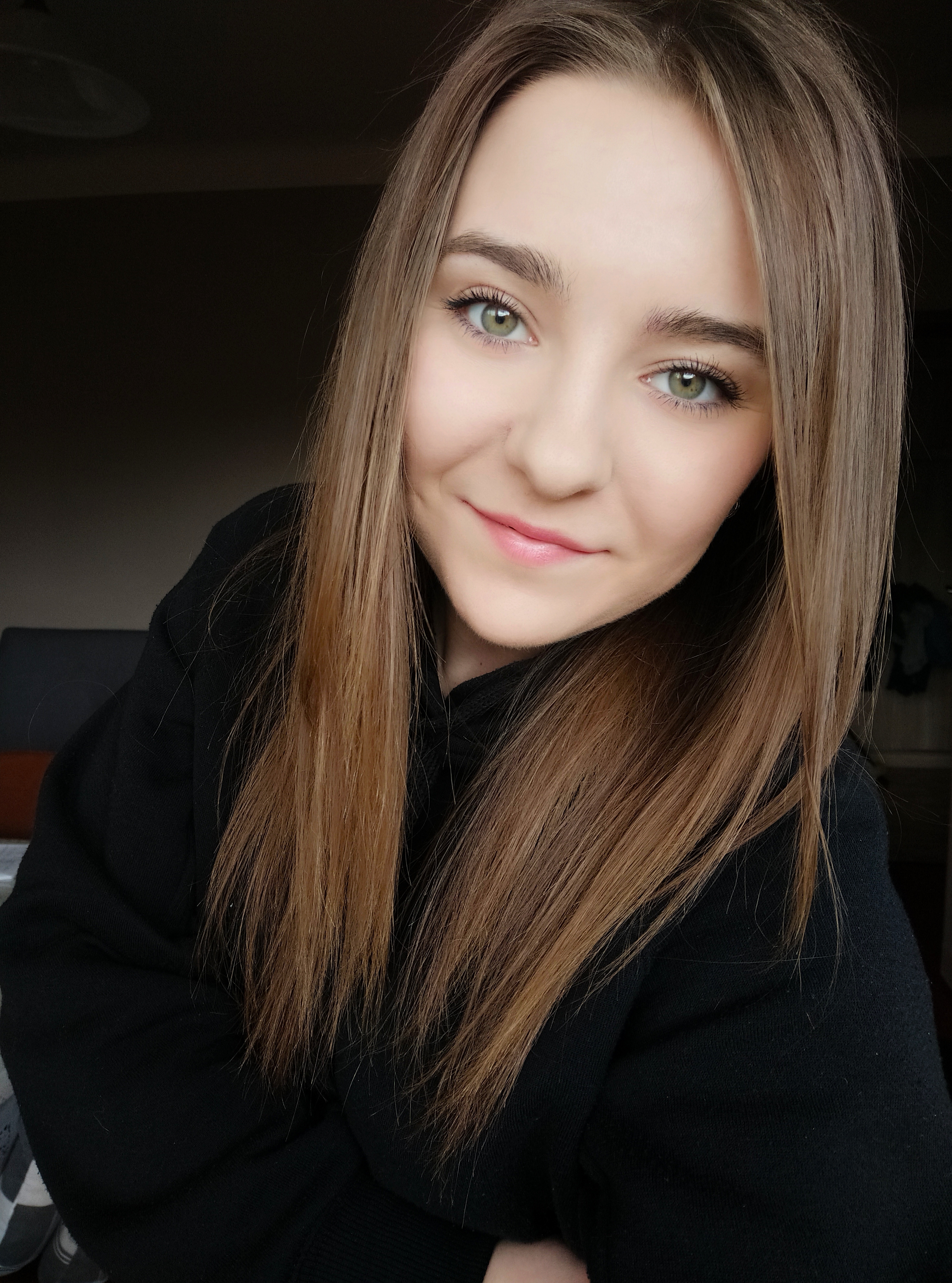 Portret uśmiechniętej młodej kobiety z długimi, brązowymi włosami i zielonymi oczami, ubrana w czarną bluzę, na rozmytym tle.