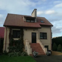 Dwupiętrowy dom z beżową elewacją, czerwoną dachówką i schodami wejściowymi, ozdobiony pnączami i kwiatami w donicach.