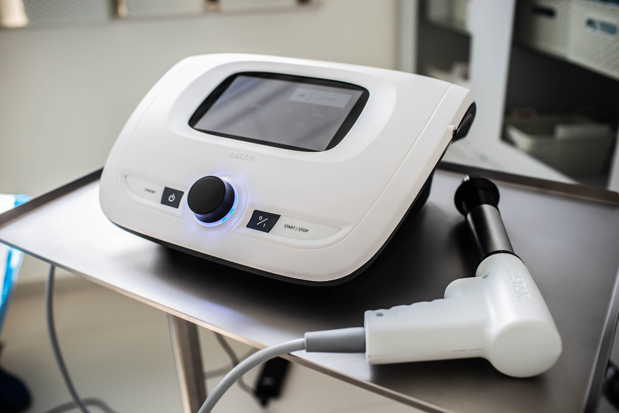 Białe urządzenie do terapii z ekranem dotykowym i aplikatorem na metalowym stoliku, widoczne przyciski i pokrętło z niebieskim podświetleniem, w tle szafka z szufladami.