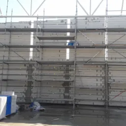 Wznoszenie ściany z bloczków Silka na rusztowaniu, widoczne palety z bloczkami i mokra posadzka w hali.