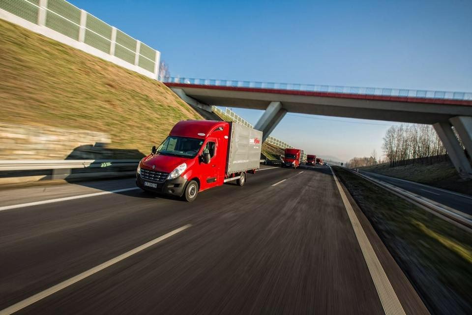 Czerwony bus dostawczy z szarą zabudową kontenerową, z logo firmy, jadący po autostradzie pod betonowym wiaduktem; w tle widoczna kolumna identycznych pojazdów.