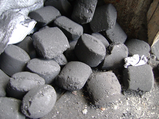 Ciemnoszare, cylindryczne bryły paliwa w worku jutowym, widoczne drobne pęknięcia i nierówności na powierzchni, biały fragment materiału między bryłami.