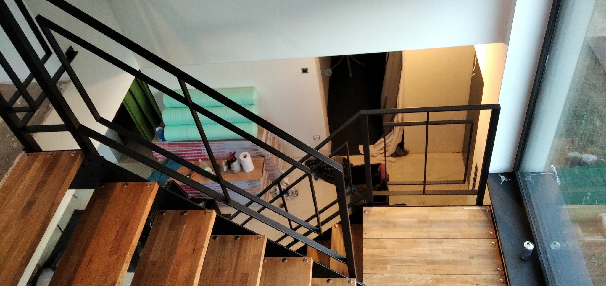 Nowoczesne schody drewniane z czarną metalową balustradą, widok z góry, w tle materiały budowlane i otwór drzwiowy.