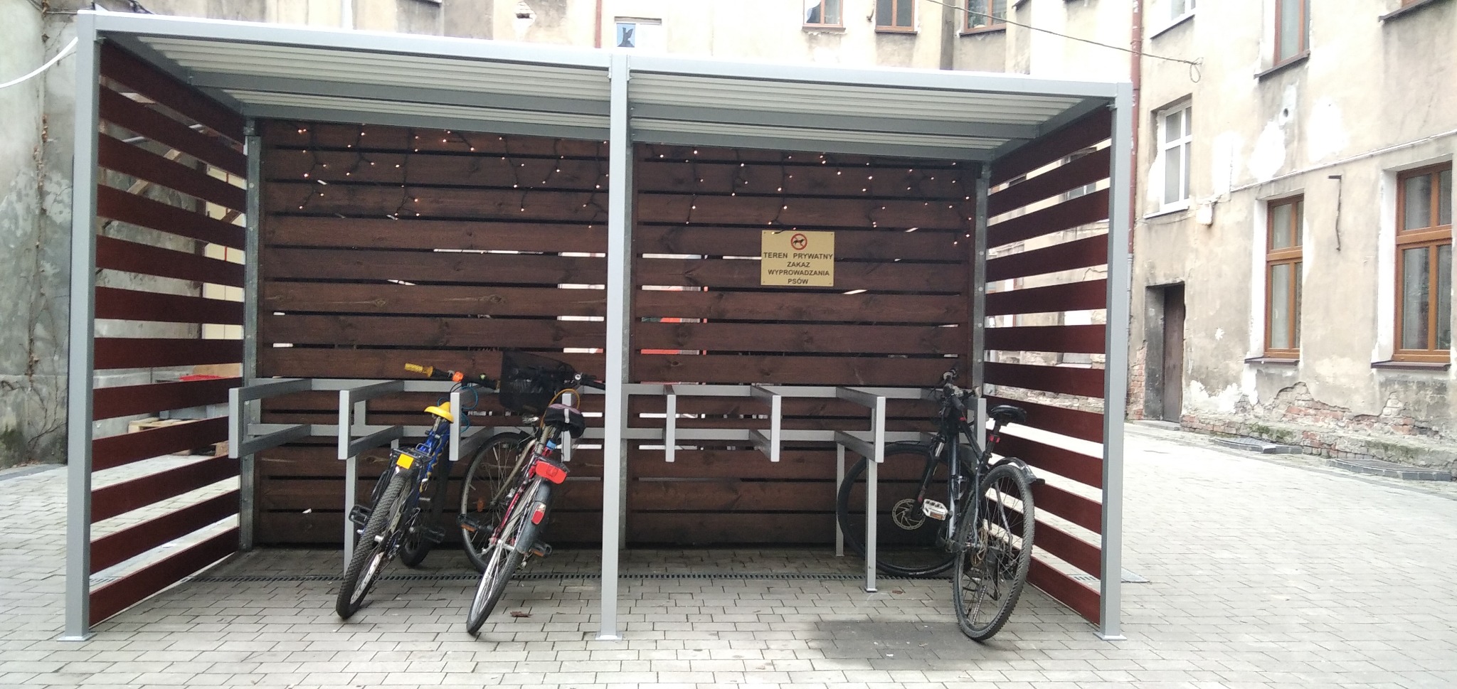 Zadaszona wiata rowerowa z drewnianymi ścianami i metalową konstrukcją, na tle starej kamienicy, z rowerami zaparkowanymi wewnątrz.