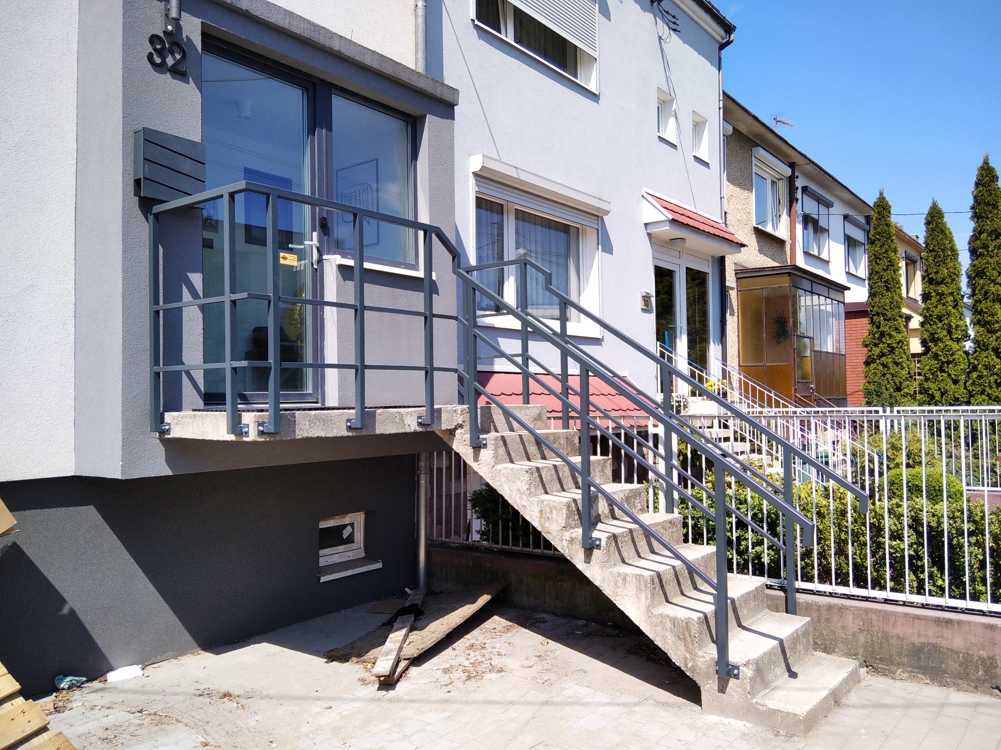 Zewnętrzne schody betonowe z szarą, metalową balustradą prowadzące do wejścia domu z oknem, widoczne elementy elewacji i otoczenie budynku.