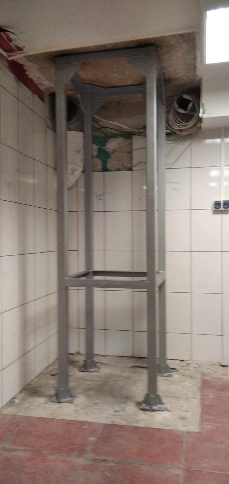 Metalowa konstrukcja wsporcza w pomieszczeniu z białymi kafelkami i czerwoną podłogą, widoczne przewody elektryczne w suficie.