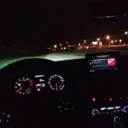 Wnętrze samochodu nocą, widok na deskę rozdzielczą z podświetlonymi zegarami i ekranem systemu multimedialnego podczas jazdy po oświetlonej ulicy.