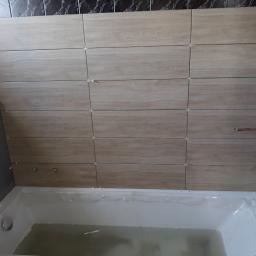 EuroMarBud - Ściana nad wanną w trakcie wykańczania, widoczne płytki imitujące drewno z krzyżykami dystansowymi, rury miedziane wystające ze ściany, wanna częściowo zakryta folią ochronną.