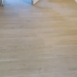 Ułożona podłoga z płytek imitujących drewno w odcieniach beżu i brązu, z widocznymi fugami w kolorze zbliżonym do płytek, prowadząca w kierunku otwartych drzwi.