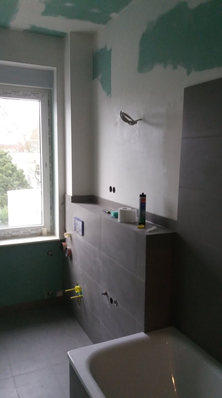 Łazienka w trakcie remontu: częściowo położone szare płytki, sufit z widocznymi śladami szpachlowania, okno z widokiem na drzewa, fragment wanny, rury instalacyjne i akcesoria budowlane na blacie.