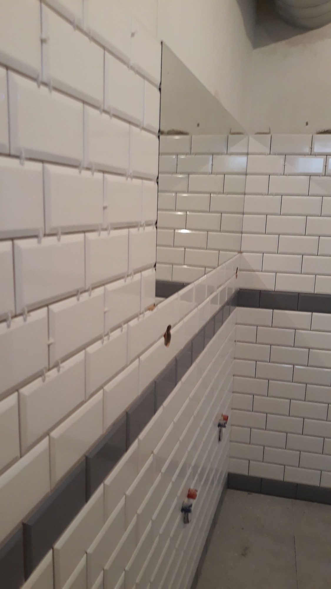 Białe, prostokątne płytki typu metro z szarym pasem dekoracyjnym, ułożone na ścianie w łazience w Poznaniu, z widocznymi krzyżykami dystansowymi i elementami instalacji wodnej.