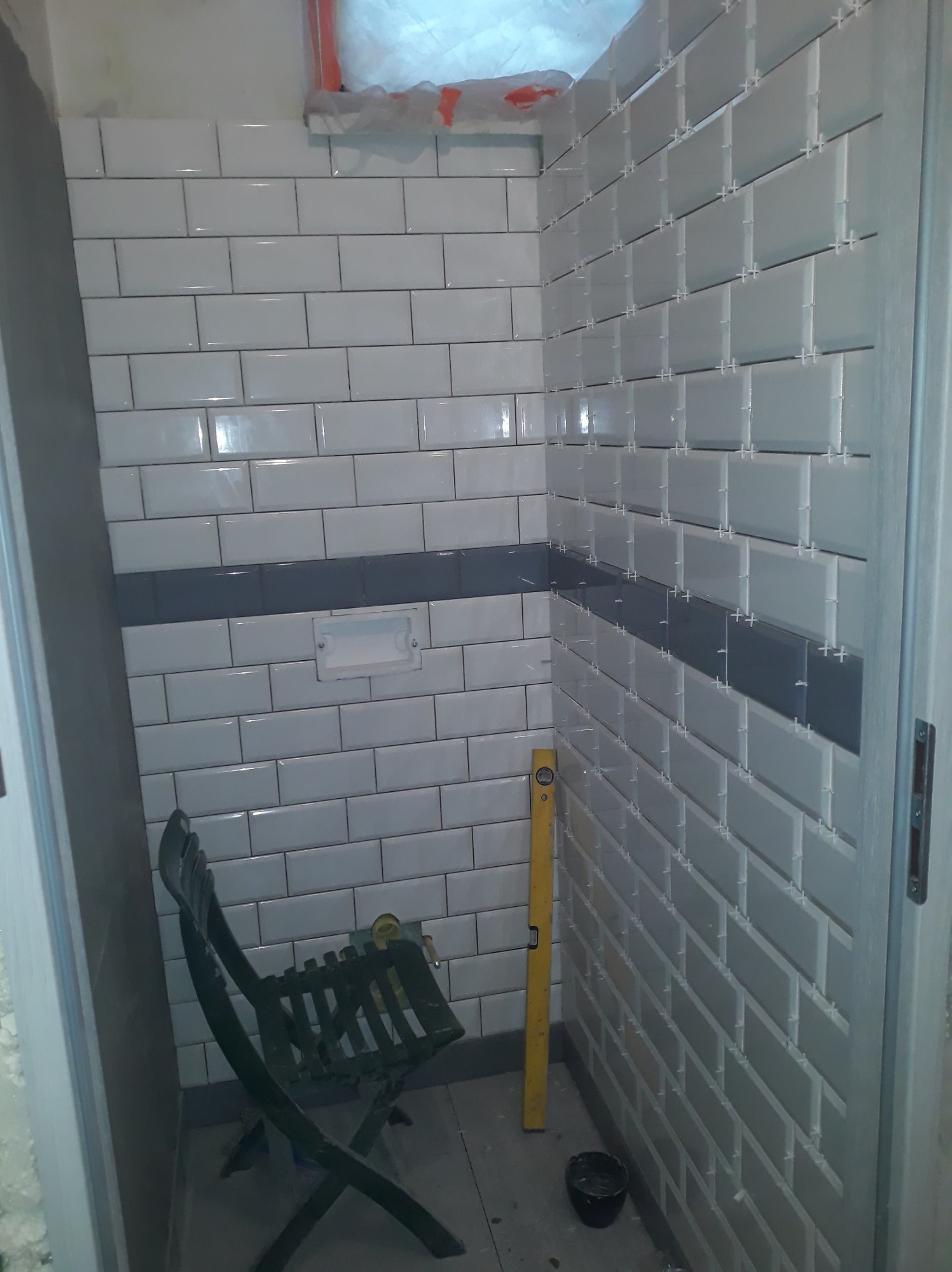 Świeżo ułożone białe płytki typu metro z szarą linią dekoracyjną, krzyżyki dystansowe widoczne, poziomica oparta o ścianę, przygotowania do fugowania w małej łazience.